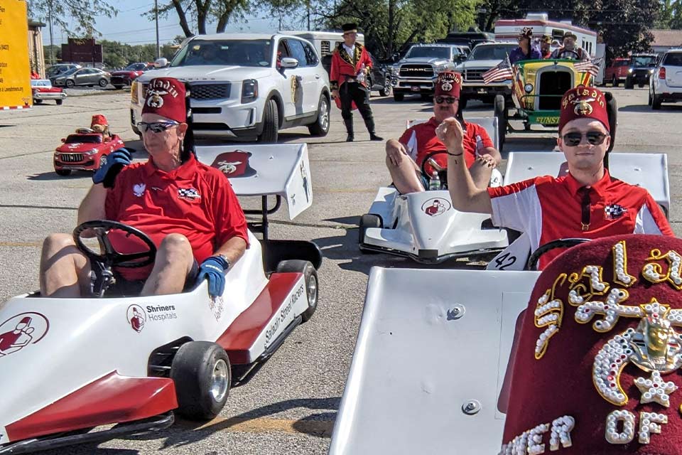three adult men wearing fezzes driving miniature race cars