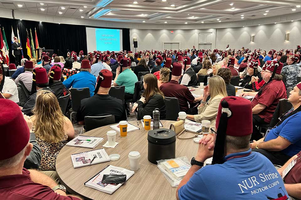 a large assembly of people wearing fezzes looking towards a screen that reads "Important Housekeeping Notes"