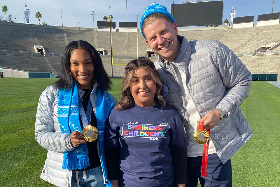 uma adolescente vestindo uma camisa que diz "Sou filha do Shriners Children's!" entre dois atletas olímpicos com suas medalhas de ouro em uma arena vazia