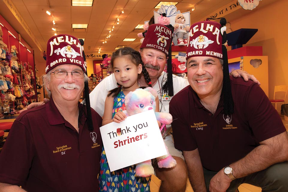 Três Shriners usando barretes vermelhos com uma garotinha segurando um bicho de pelúcia e uma placa que diz: Obrigado Shriners;