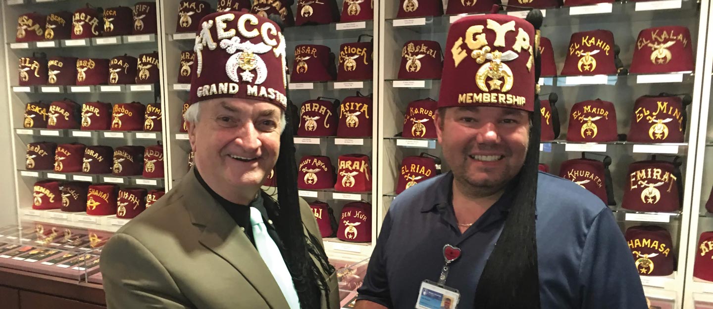 A Mecca Shriner and Egypt Shriner, both wearing their fez, standing in front of a wall of fezzes